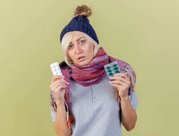 De bleke jonge blonde zieke slavische vrouw die de wintermuts en sjaal draagt houdt verpakkingen van medische pillen die op olijfgroene muur met exemplaarruimte worden geïsoleerd