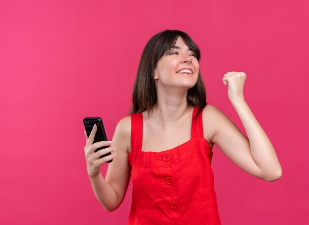 De blije jonge Kaukasische telefoon van de meisjesholding en het houden van vuist omhoog kijkend naar de kant op geïsoleerde roze achtergrond met exemplaarruimte