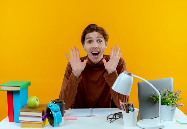 De blije jonge zitting van de studentenjongen aan bureau met de hand van schoolhulpmiddelen rond gezicht