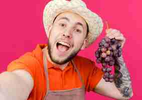 Gratis foto de blije mannelijke tuinman die het tuinieren hoed draagt houdt druiven die beweren camera vast te houden
