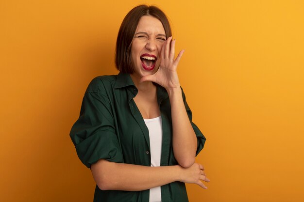 De blije mooie vrouw houdt hand dicht bij mond die iemand roept die op oranje muur wordt geïsoleerd