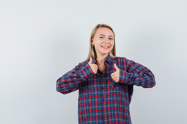 De blonde vrouw die duimen met beide toont dient gecontroleerd overhemd in en kijkt gelukkig, vooraanzicht.