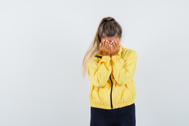 De blonde vrouw die gezicht behandelt met dient gele bomberjack en zwarte broek in en op zoek gekweld