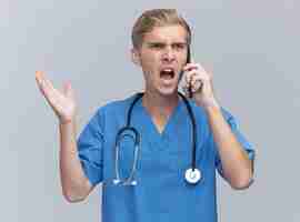 Gratis foto de boze jonge mannelijke arts die artsenuniform met stethoscoop draagt spreekt over telefoon die die hand spreidt op witte muur wordt geïsoleerd