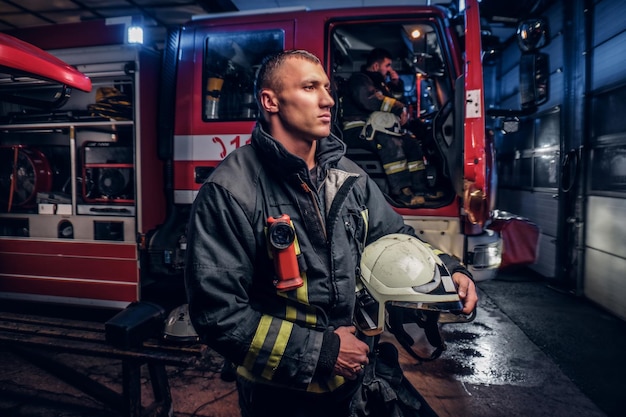 De brandweer kwam 's nachts ter plaatse. Knappe brandweerman die een beschermend uniform draagt met een zaklamp erbij, een helm vasthoudt en opzij kijkt