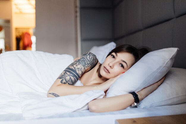 De donkerbruine vrouw met tatoegering legt in het bed op het hoofdkussen in de ochtend in manier modern appartement