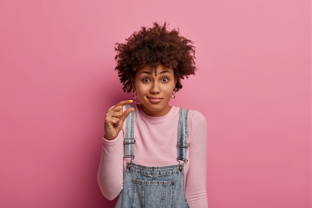 De foto van een verwarde vrouw met een donkere huid toont een klein formaat met vingers, vertelt over een verlaagde prijs, toont een kleine afmeting, vraagt om een klein bod, heeft een teleurgestelde uitdrukking, staat binnen.