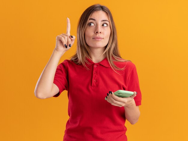 De geïmponeerde jonge mooie vrouw houdt telefoon vast en wijst omhoog geïsoleerd op oranje muur