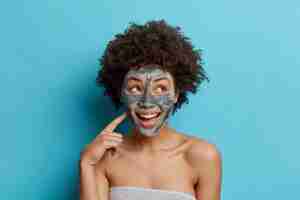 Gratis foto de gelukkige etnisch gekrulde vrouw glimlacht aangenaam past gezichtskleimasker wil er mooi uit verpakt in zachte handdoek die over blauwe studiomuur wordt geïsoleerd.