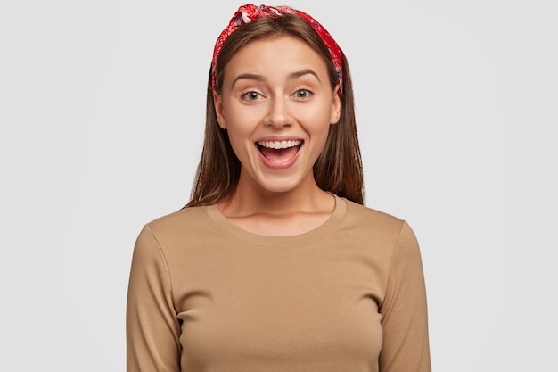 De gelukkige Europese vrouw met groene ogen met tevreden uitdrukking, houdt mond open, draagt rode bandana