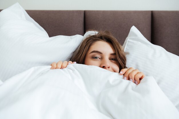 De gelukkige jonge dame ligt thuis in bed onder een deken.