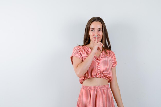 De gelukkige jonge vrouw toont een stil gebaar door haar wijsvinger op de lippen op een witte achtergrond te houden