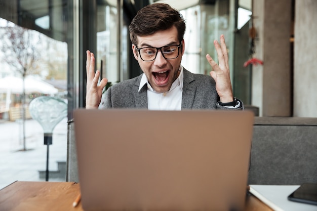 De geschokte gillende zakenman in oogglazen die door de lijst in koffie zitten en verheugt zich terwijl het bekijken laptop computer