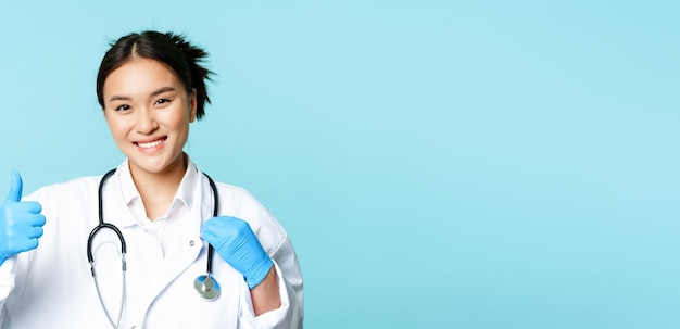 De glimlachende aziatische arts van de vrouwentherapeut met stethoscoop toont duimen die zich over blauwe backgrou bevinden