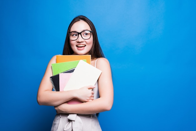 De glimlachende Aziatische boeken en het dossier van de studenteholding, hogeschool of schoolstudent en onderwijsconcept dat op blauwe muur met exemplaarruimte wordt geïsoleerd.