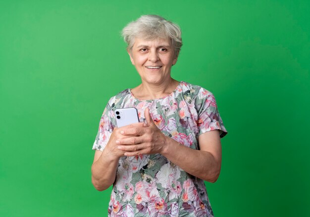 De glimlachende bejaarde houdt telefoon die op groene muur wordt geïsoleerd