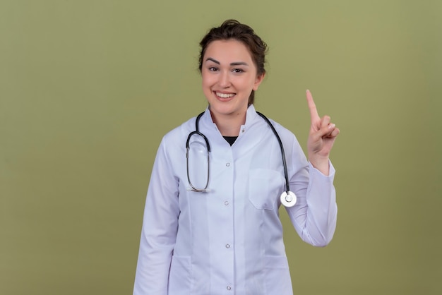 De glimlachende jonge arts die medische toga draagt die stethoscoop draagt, toont één op groene muur