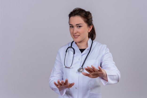 De glimlachende jonge arts die medische toga draagt die stethoscoop draagt, toont grootte op witte muur