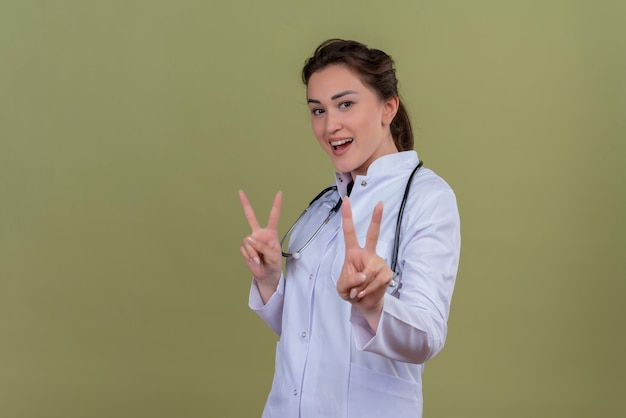 De glimlachende jonge arts die medische toga draagt die stethoscoop draagt, toont vredesgebaar op groene muur