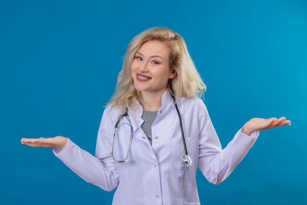 De glimlachende jonge arts die stethoscoop in medische toga draagt spreidt handen uit op blauwe muur