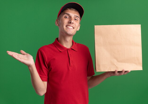 De glimlachende jonge blonde bezorger houdt de hand open en houdt papierpakket vast dat op groene muur met exemplaarruimte wordt geïsoleerd