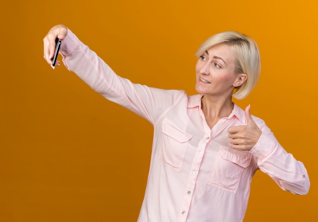 De glimlachende jonge blonde slavische vrouw neemt een selfie haar duim omhoog geïsoleerd op oranje