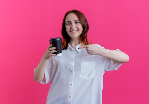 De glimlachende jonge holdings van het roodharigemeisje en wijst op kop van koffie die op roze wordt geïsoleerd