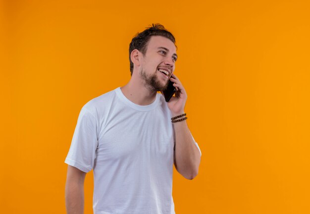 De glimlachende jonge kerel die wit t-shirt draagt spreekt over telefoon op geïsoleerde oranje achtergrond