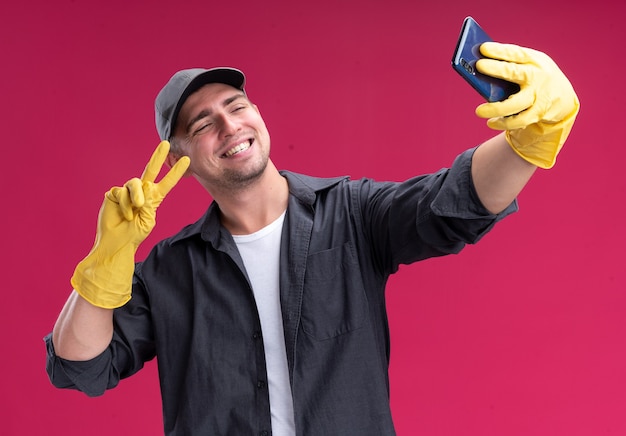 De glimlachende jonge knappe schoonmakende kerel die t-shirt en pet met handschoenen draagt, neemt een selfie die vredesgebaar toont dat op roze muur wordt geïsoleerd