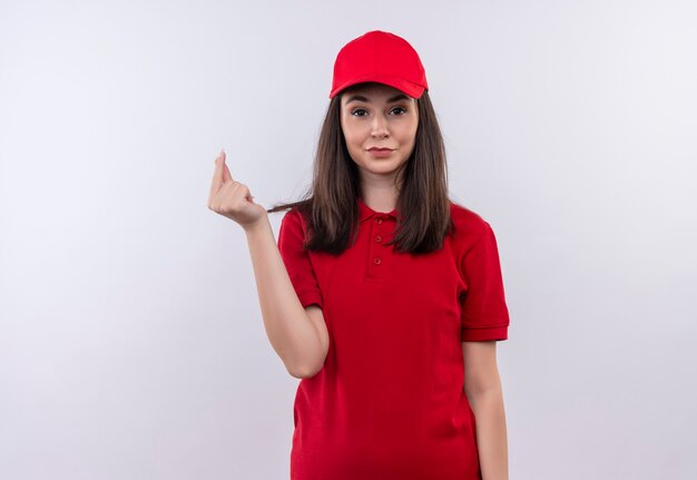 De glimlachende jonge leveringsvrouw die een rode t-shirt in een rode pet draagt, vraagt om een fooi op geïsoleerde witte muur