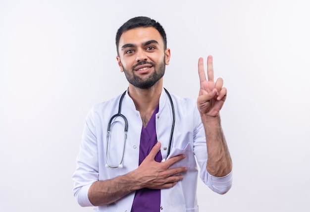 De glimlachende jonge mannelijke arts die stethoscoop medische toga draagt die vredesgebaar toont, legde zijn hand op hart op geïsoleerde witte achtergrond