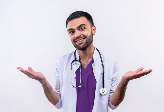 De glimlachende jonge mannelijke arts die stethoscoop medische toga draagt, spreidt handen op geïsoleerde witte achtergrond uit