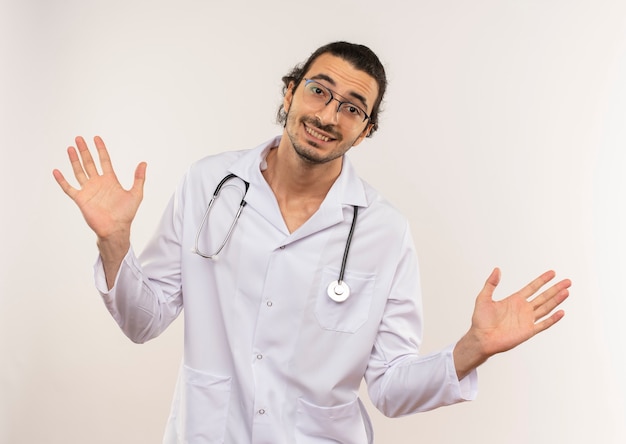 De glimlachende jonge mannelijke arts met optische bril die wit gewaad met stethoscoop draagt spreidt handen op geïsoleerde witte muur met exemplaarruimte uit