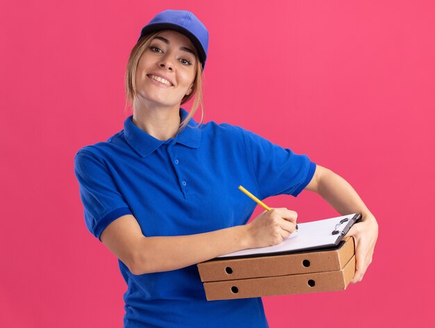 De glimlachende jonge mooie bezorger in uniform houdt pizzadozen en schrijft op klembord met pen die op roze muur wordt geïsoleerd