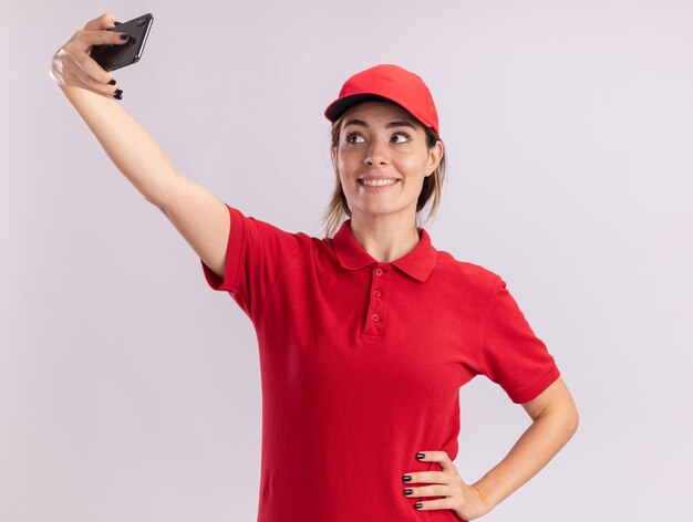 De glimlachende jonge mooie leveringsvrouw in uniform houdt en kijkt naar telefoon die selfie neemt die op witte muur wordt geïsoleerd