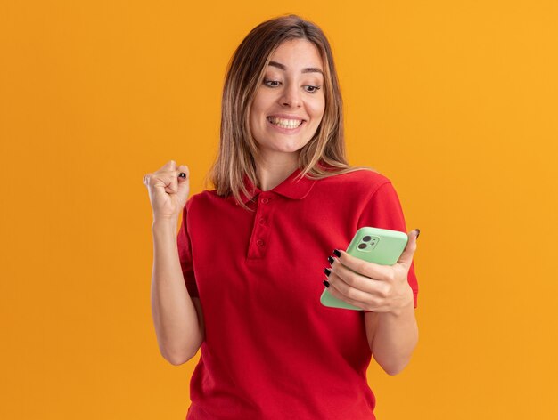 De glimlachende jonge mooie vrouw houdt vuist houdend en bekijkt telefoon die op oranje muur wordt geïsoleerd