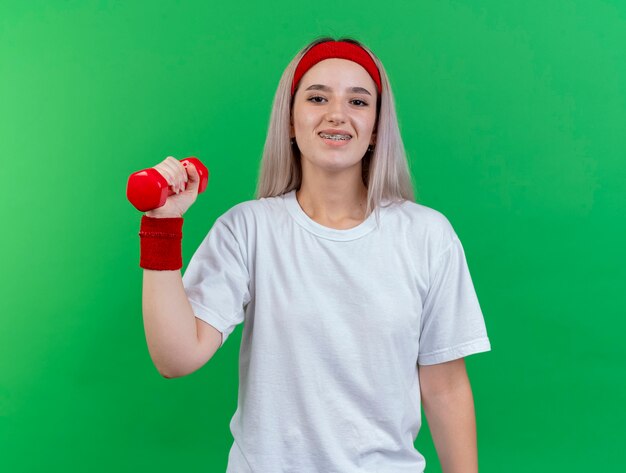 De glimlachende jonge sportieve vrouw met bretels die hoofdband en polsbandjes dragen houdt domoor die op groene muur wordt geïsoleerd