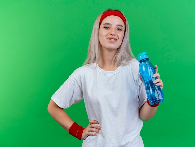 De glimlachende jonge sportieve vrouw met bretels die hoofdband en polsbandjes dragen houdt waterfles en legt hand op taille die op groene muur wordt geïsoleerd