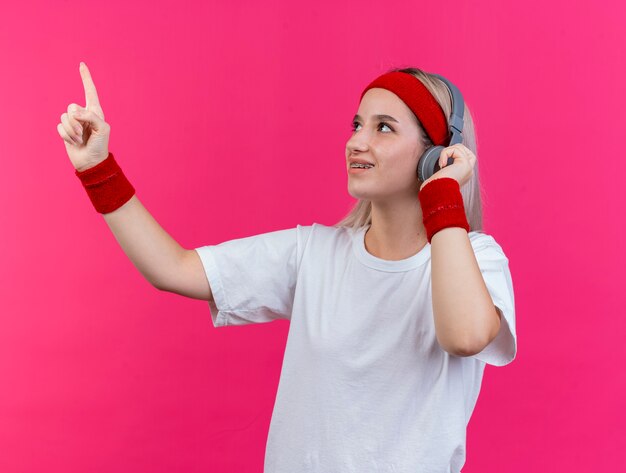 De glimlachende jonge sportieve vrouw met steunen op hoofdtelefoons die hoofdband en polsbandjes dragen kijkt en wijst naar kant geïsoleerd op roze muur