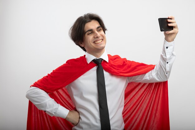 De glimlachende jonge superheldenkerel die stropdas draagt, neemt aselfie en zet hand op heup die op witte achtergrond wordt geïsoleerd