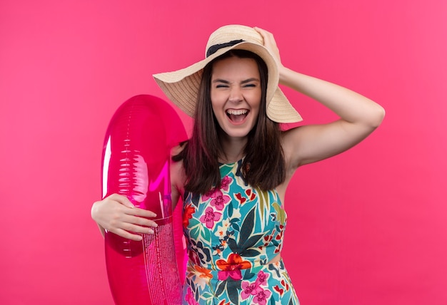 De glimlachende jonge vrouw die hoed draagt die zwemt ring houdt en rotsteken op geïsoleerde roze muur toont