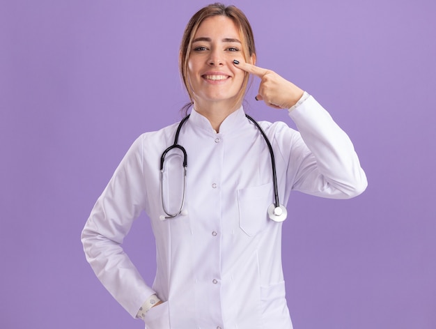 De glimlachende jonge vrouwelijke arts die medisch kleed met stethoscoop draagt ?? richt op oog dat op purpere muur wordt geïsoleerd