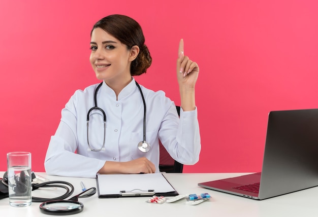 De glimlachende jonge vrouwelijke arts die medische kleed met stethoscoop draagt die aan bureau werkt op computer met medische hulpmiddelen wijst met exemplaarruimte omhoog