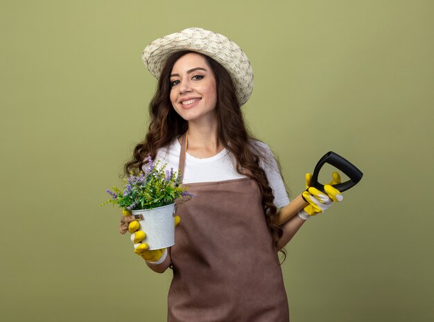 De glimlachende jonge vrouwelijke tuinman in eenvormig die tuinierende hoed en handschoenen draagt houdt bloempot en schop achter rug geïsoleerd op olijfgroene muur