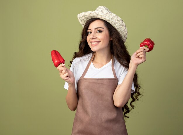 De glimlachende jonge vrouwelijke tuinman in uniform die het tuinieren hoed draagt houdt rode paprika's die op olijfgroene muur worden geïsoleerd