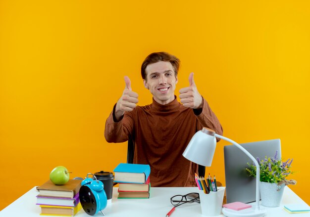 De glimlachende jonge zitting van de studentenjongen aan bureau met schoolhulpmiddelen zijn duimen omhoog geïsoleerd op gele muur