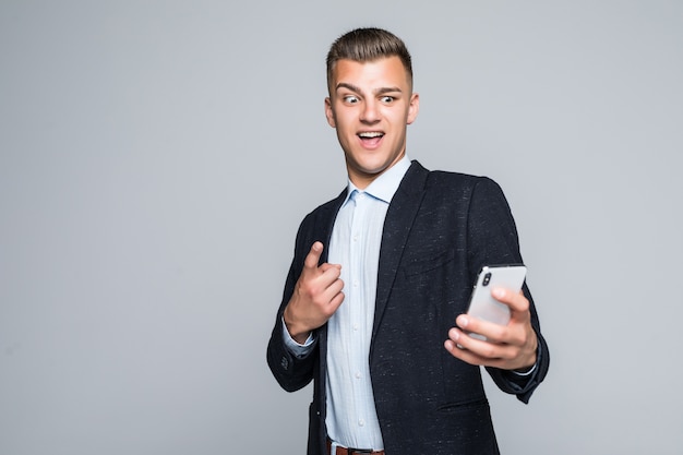 De glimlachende jongeman heeft een videogesprek op een telefoon gekleed in donker jasje in studio die op grijze muur wordt geïsoleerd