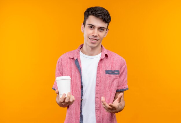 De glimlachende Kaukasische jonge kerel die roze overhemd draagt die kop van koffie houdt stak hand op geïsoleerde oranje achtergrond uit