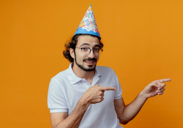 De glimlachende knappe mens die glazen en verjaardag glb-punten draagt aan kant die op oranje achtergrond met exemplaarruimte wordt geïsoleerd