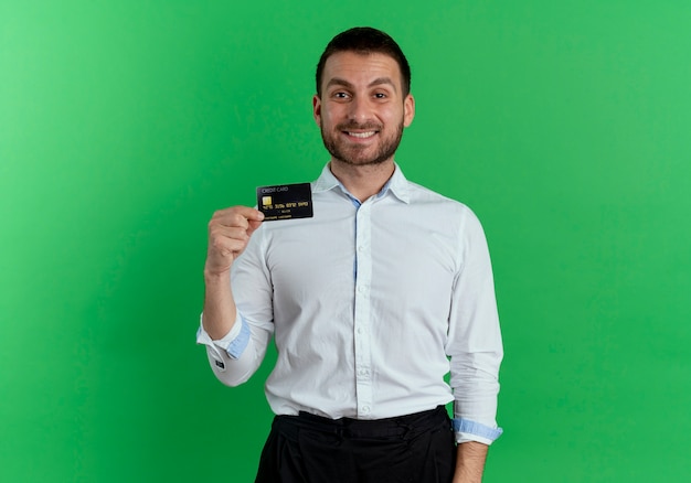 De glimlachende knappe mens houdt creditcard die op groene muur wordt geïsoleerd